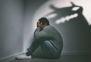 Shot of a young man sitting in the corner of a dark room with a scary figure on the wall.