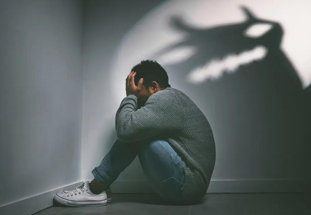 How Trauma Shows Up In Men Who Are Good At Hiding Their Feelings Shot of a young man sitting in the corner of a dark room with a scary figure on the wall.