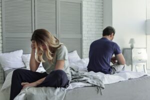 Couple sitting apart on a bed after infidelity with the woman holding her head in distress while the man faces away in emotional distance
