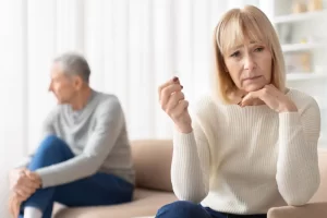 Older woman looking thoughtfully at a wedding ring while an older man sits apart in the background suggesting marital separation or divorce